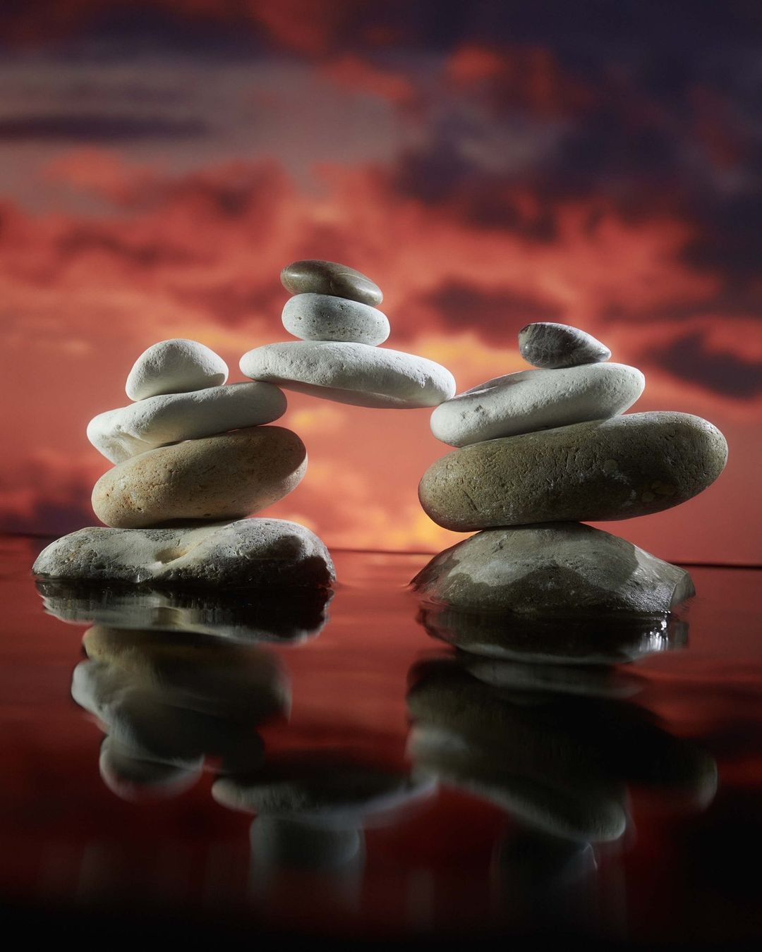 Balanced rock towers create an archway over reflecting water at sunset, set against a dramatic cloudy sky filled with oranges and purples.