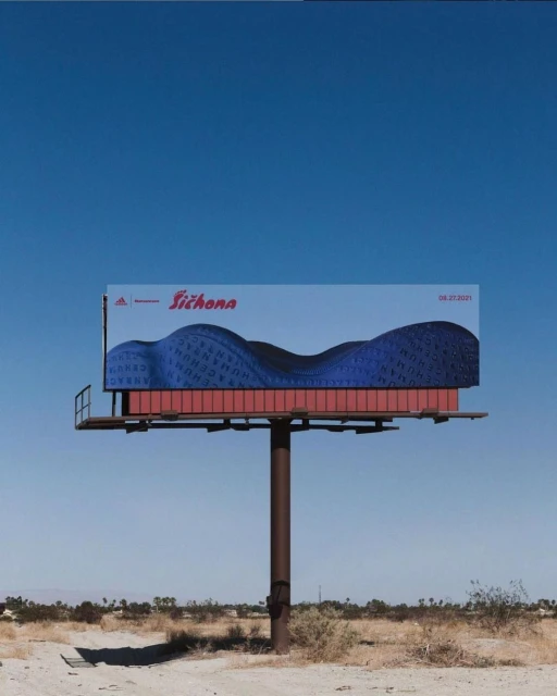 Outdoor billboard featuring an abstract design with blue hues and the Adidas logo against a clear blue sky and desert landscape.