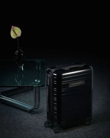 Black rolling suitcase alongside a glass table with a flower in a vase in a dark setting creates a sophisticated travel lifestyle scene.