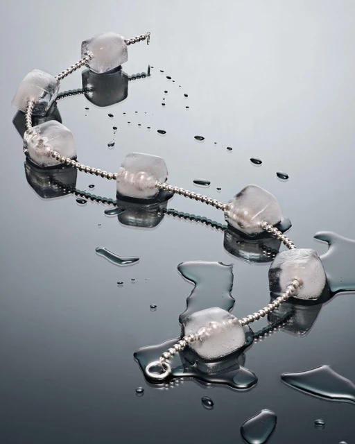 A necklace made of ice cubes connected by a silver chain, laying on a wet, reflective surface, suggesting a fusion of jewelry and nature.