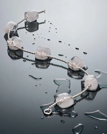 A necklace made of ice cubes connected by a silver chain, laying on a wet, reflective surface, suggesting a fusion of jewelry and nature.