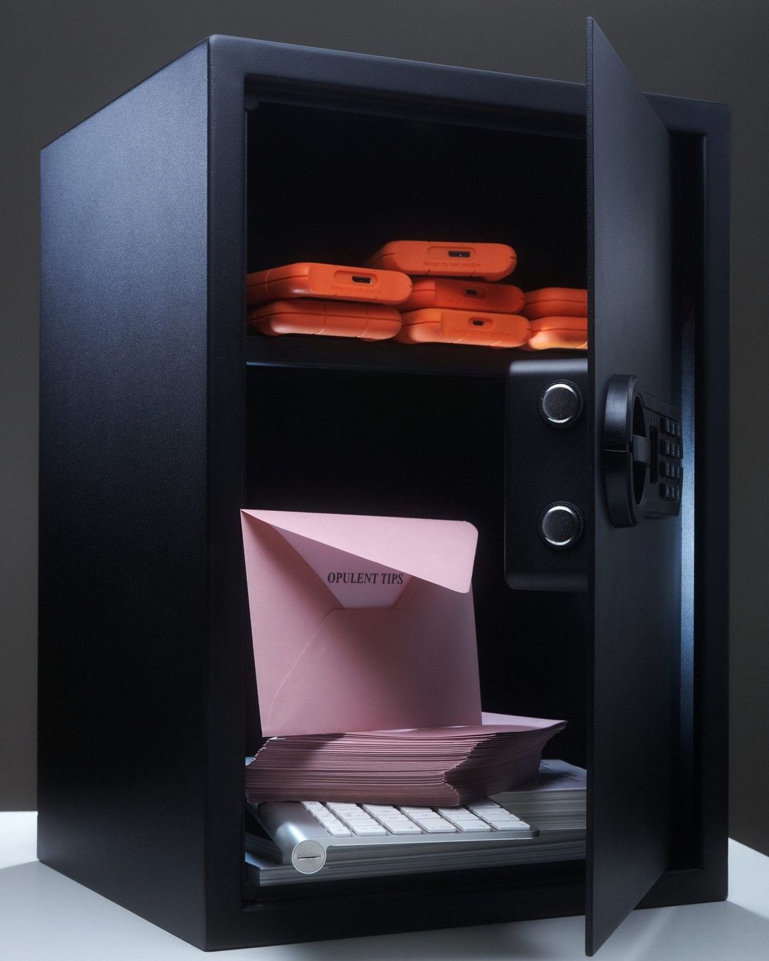 Open black safe filled with stacks of pink envelopes, orange hard drives, and a white computer keyboard, placed on a white surface.