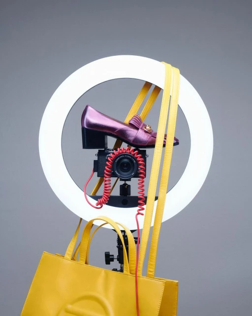 Fashion still life with a pair of metallic magenta loafers, a yellow leather tote bag, a camera, and a ring light, studio shot.