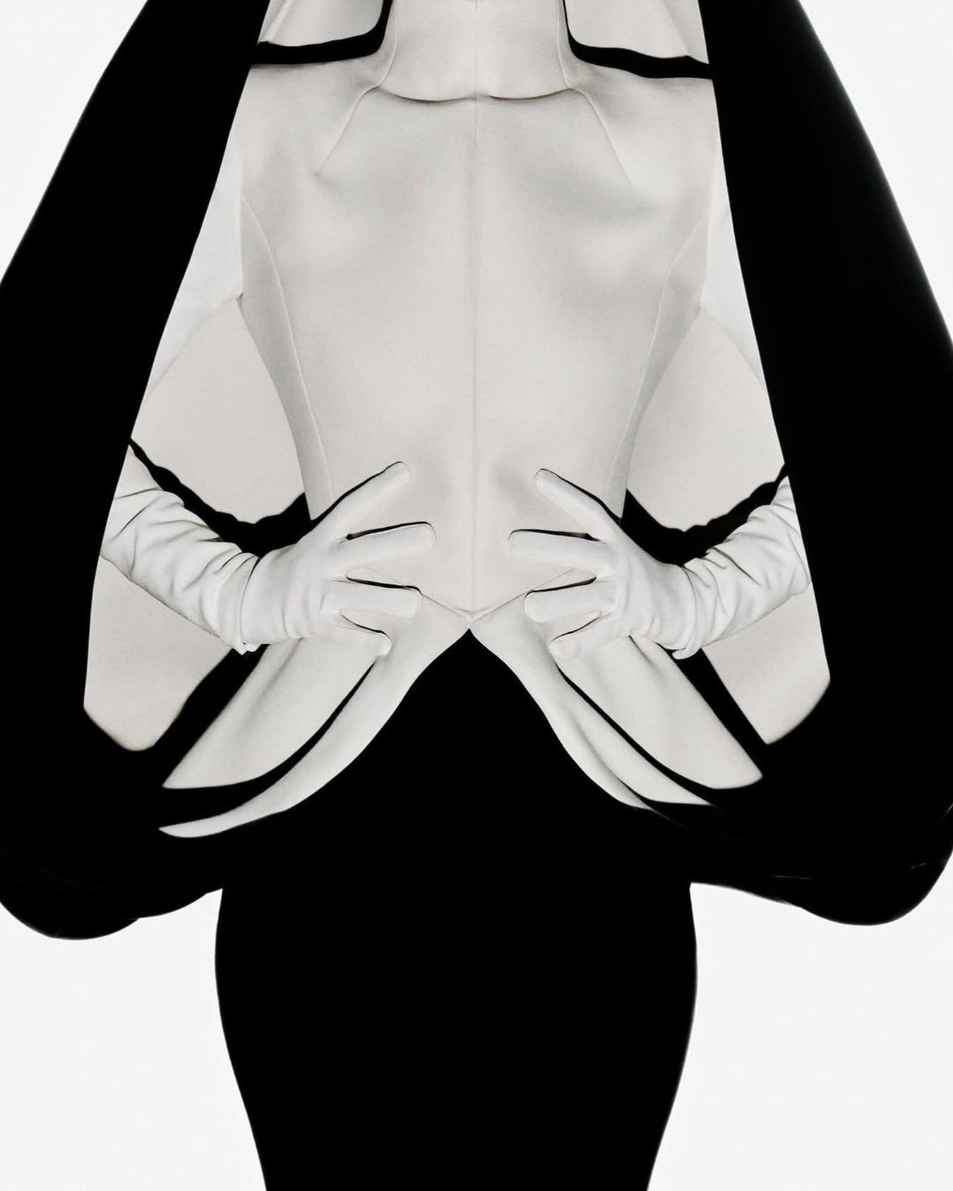 Elegant upper-body fashion portrait with a model wearing a white jacket, gloves, and a black dress against a bright background.