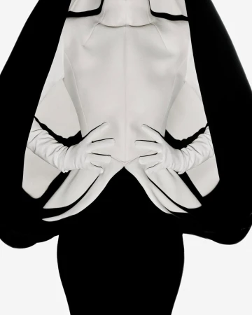 Elegant upper-body fashion portrait with a model wearing a white jacket, gloves, and a black dress against a bright background.