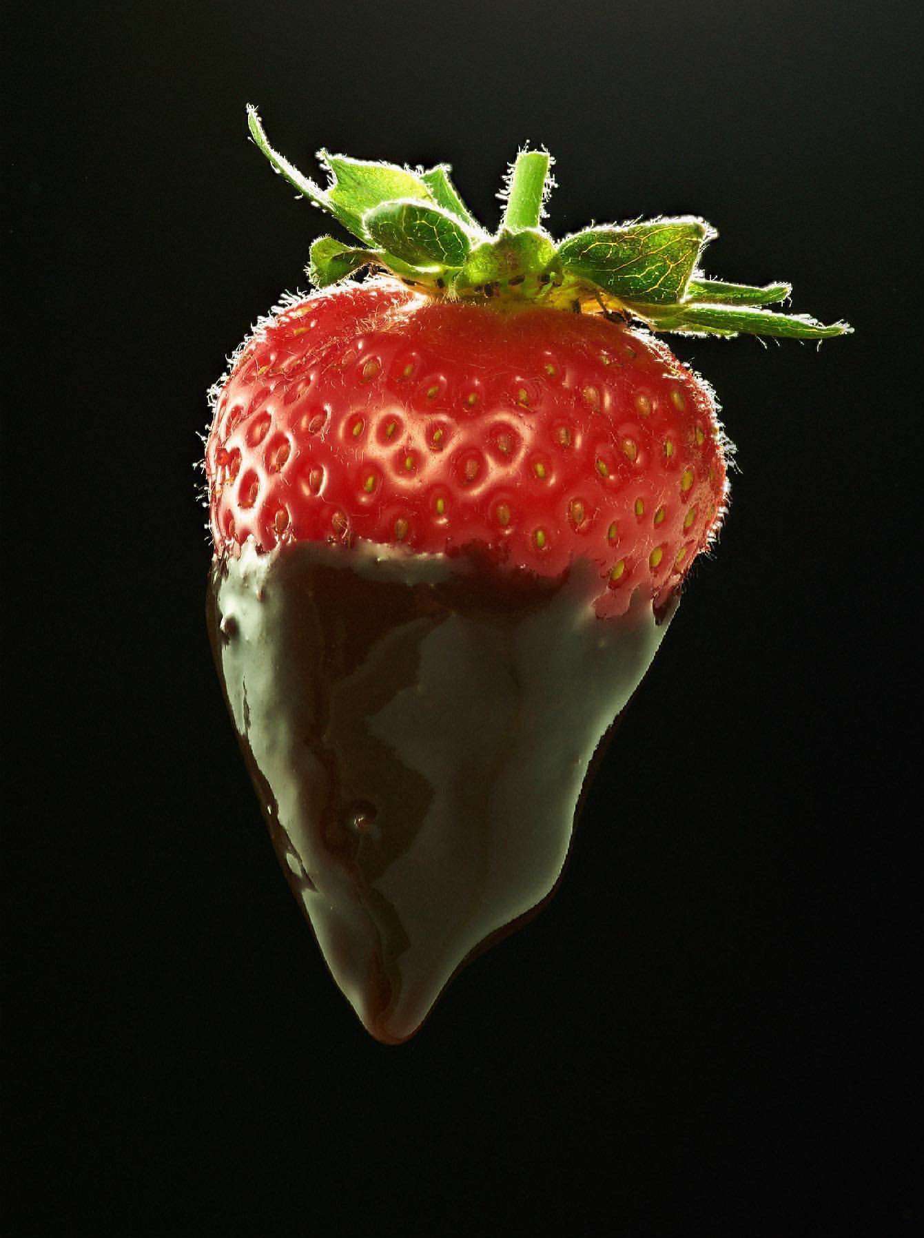 A single strawberry, partially dipped in dark chocolate, stands against a stark black background, capturing its juicy texture.