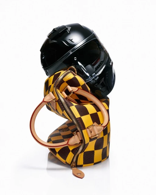 Still life of a black motorcycle helmet balanced on a checkered yellow and brown handbag against a bright white background.
