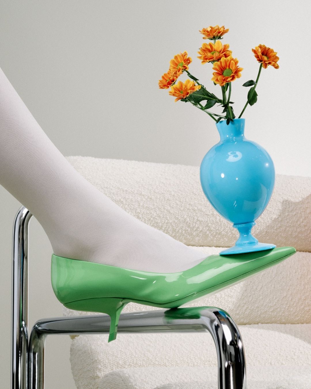 Still life of a green high-heeled shoe with a turquoise vase of orange flowers, resting on a chrome chair with a white cushion.
