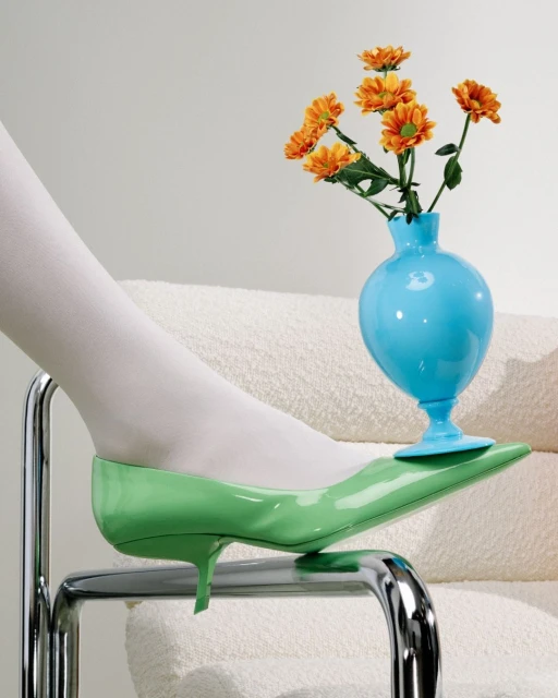 Still life of a green high-heeled shoe with a turquoise vase of orange flowers, resting on a chrome chair with a white cushion.