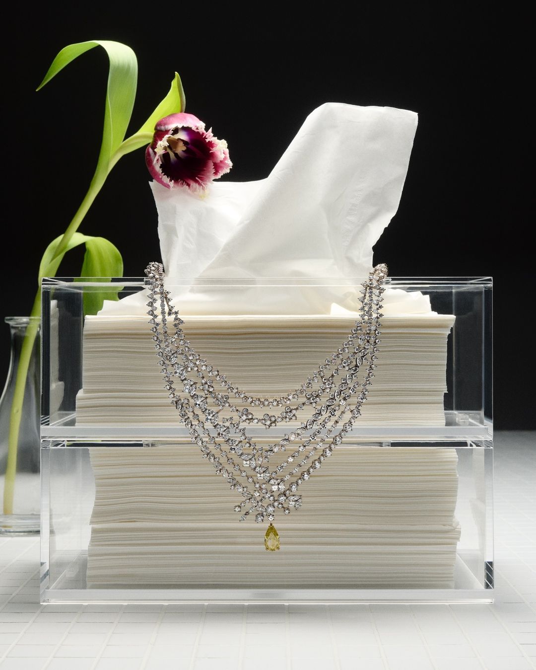 Elegant diamond necklace draped over a clear acrylic tissue box with white tissues and a purple tulip against a black background.