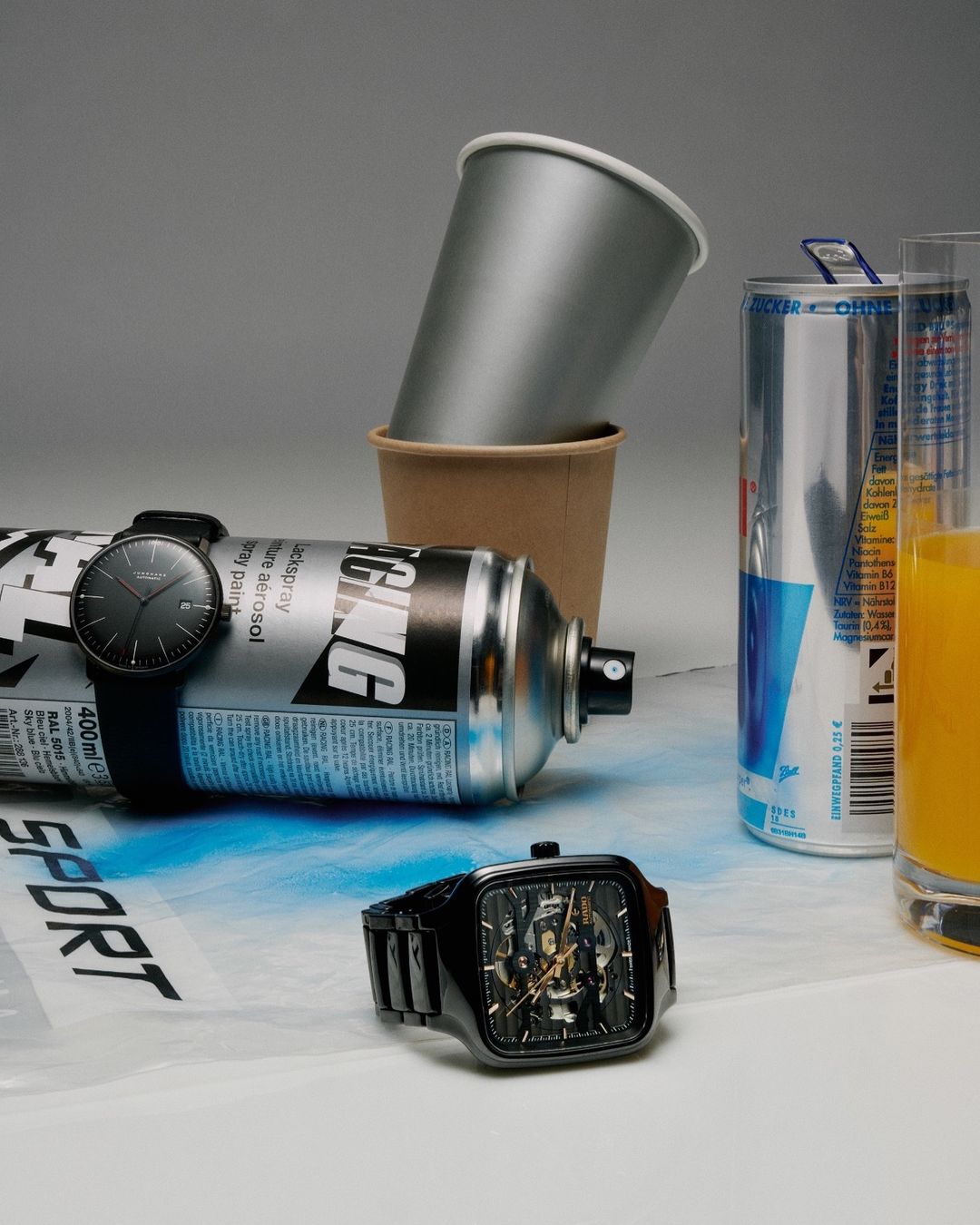 Still life composition featuring two wristwatches, spray paint can, drinking glasses and an aluminum can against a neutral grey backdrop.