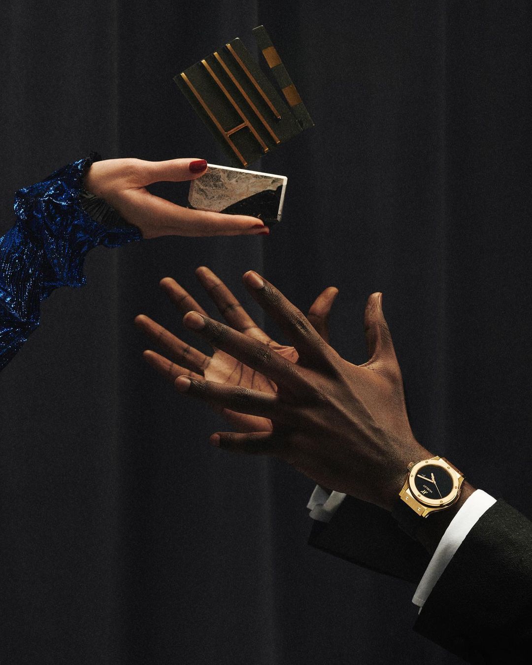Still life shot of a person's hands with a luxury watch and a person's hands holding a cosmetic product, against a dark backdrop.