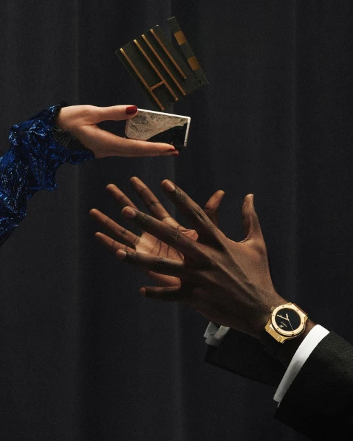 Still life shot of a person's hands with a luxury watch and a person's hands holding a cosmetic product, against a dark backdrop.