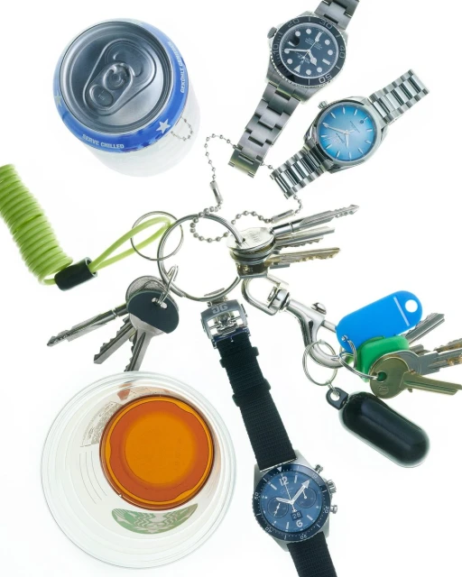 Overhead shot of a keychain with multiple keys, three wristwatches, a can, and a glass of liquid against a bright white background.