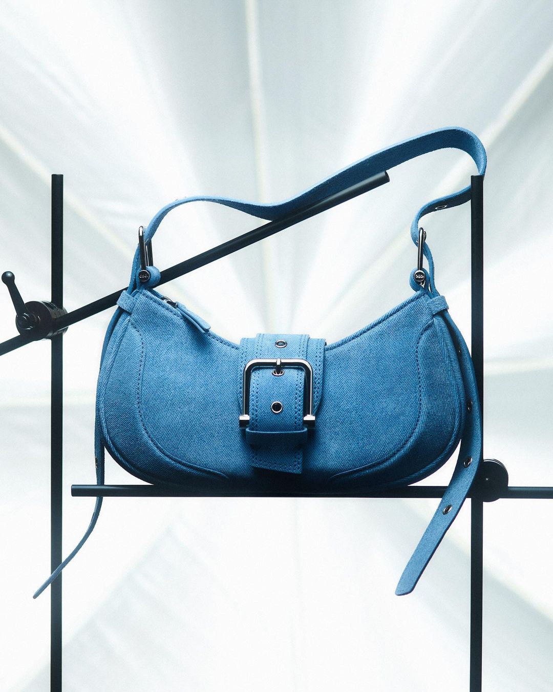 Stylish blue suede purse on black metal stand with a bright background. The handbag features a buckle closure.