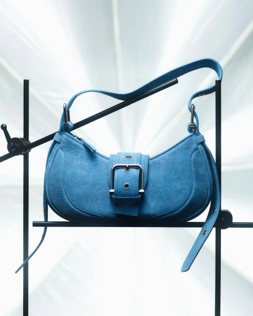 Stylish blue suede purse on black metal stand with a bright background. The handbag features a buckle closure.