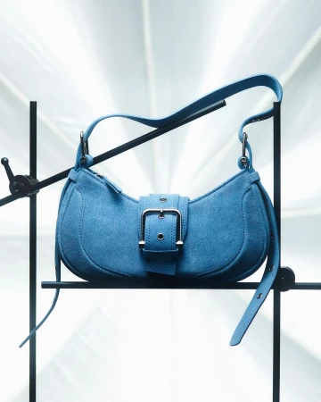 Stylish blue suede purse on black metal stand with a bright background. The handbag features a buckle closure.