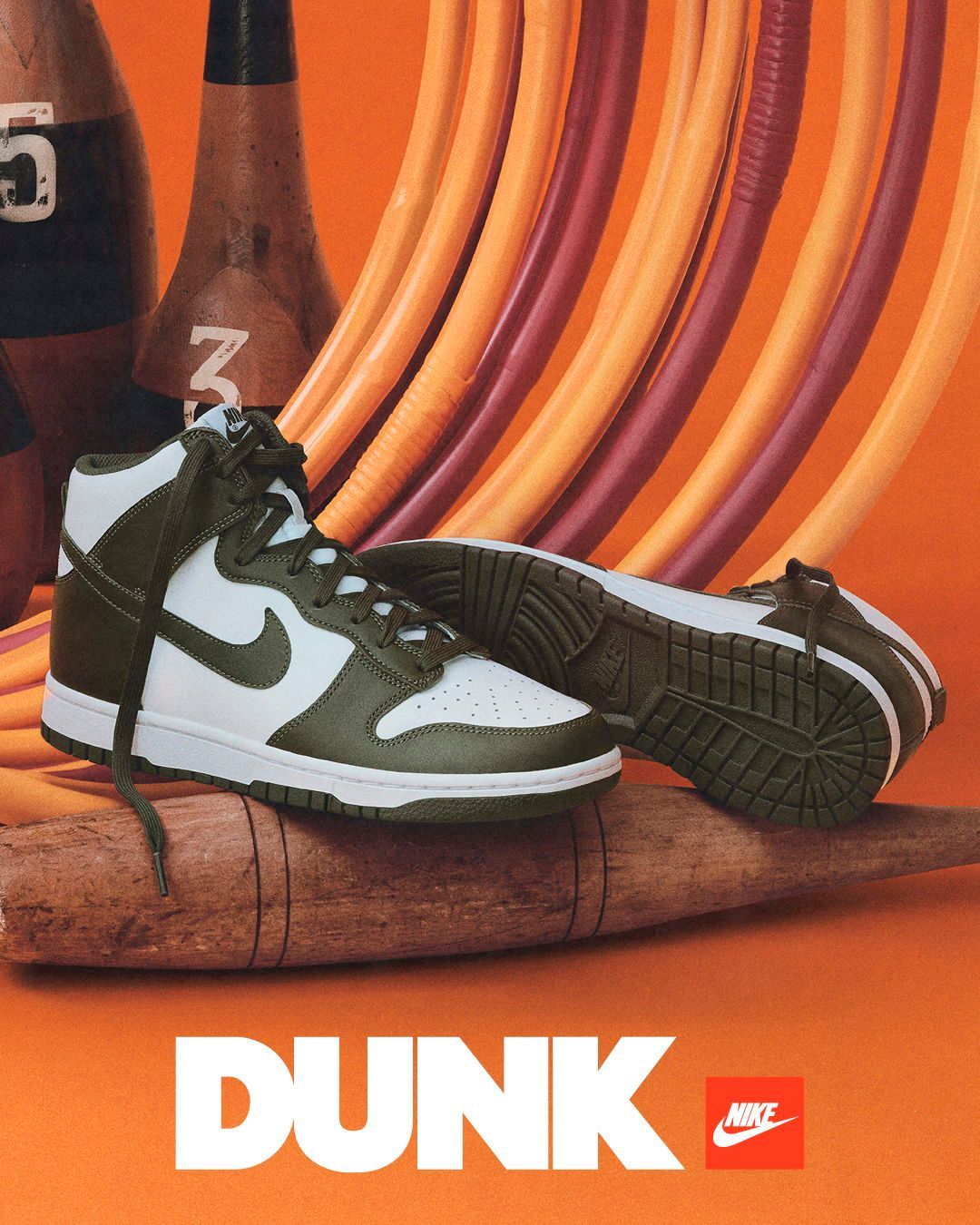 Olive green and white Nike Dunk high-top sneakers are displayed on a wooden log against an orange background with bowling pins.