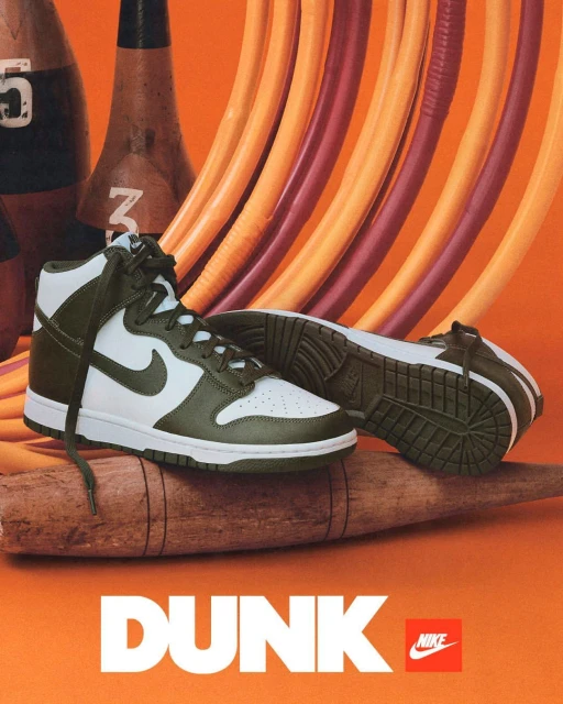 Olive green and white Nike Dunk high-top sneakers are displayed on a wooden log against an orange background with bowling pins.