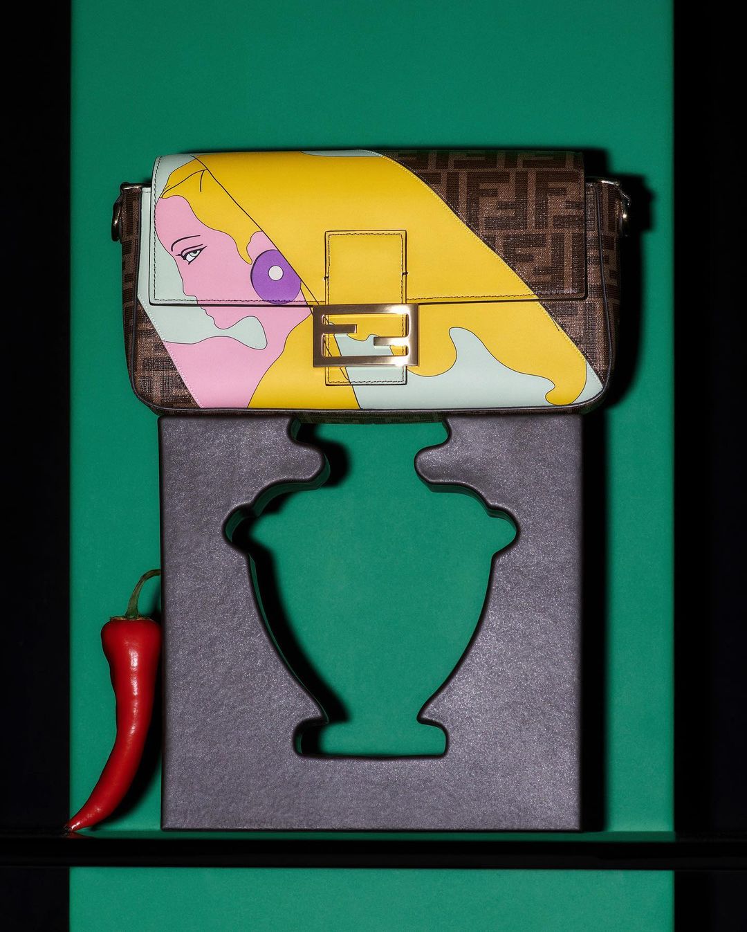 Designer purse with a graphic of a woman's face in yellow, pink, and teal, rests on a carved stand with a pepper beside it.