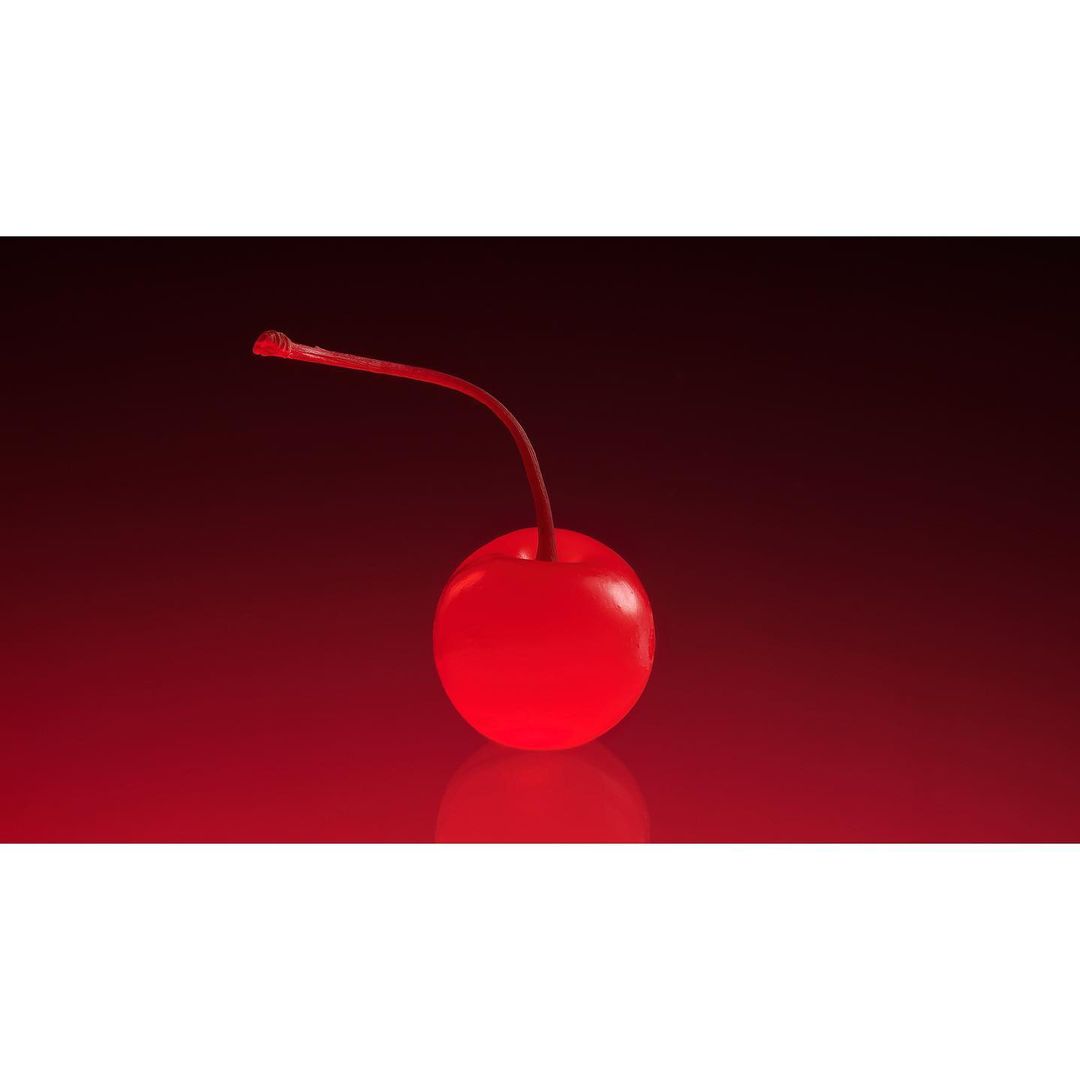 Close-up studio shot of a shiny red maraschino cherry with its stem against a dark red gradient background with a reflective surface.