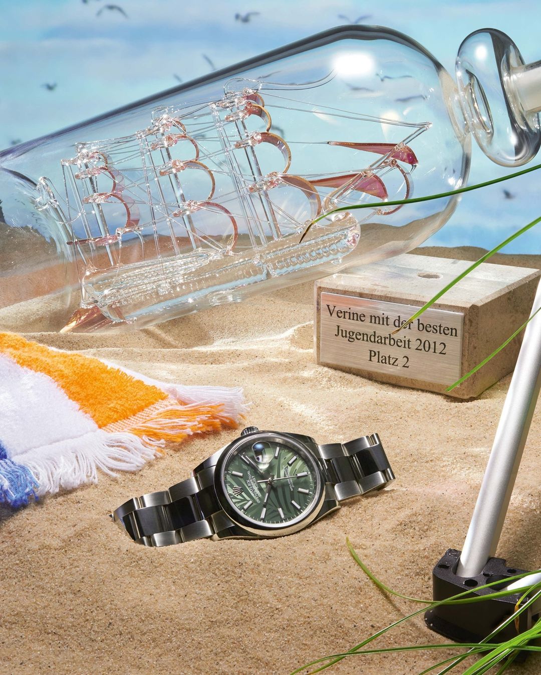 Wristwatch on sand next to ship in glass bottle with beach towel, grass, and blue sky in outdoor summer setting. Still life.