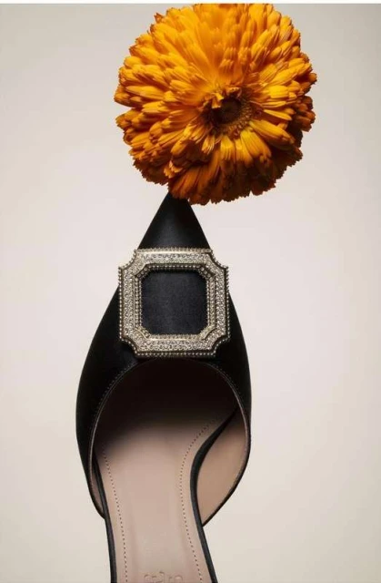 Close-up studio shot of a black satin pump with jeweled buckle, styled with an orange marigold blossom against a beige background.