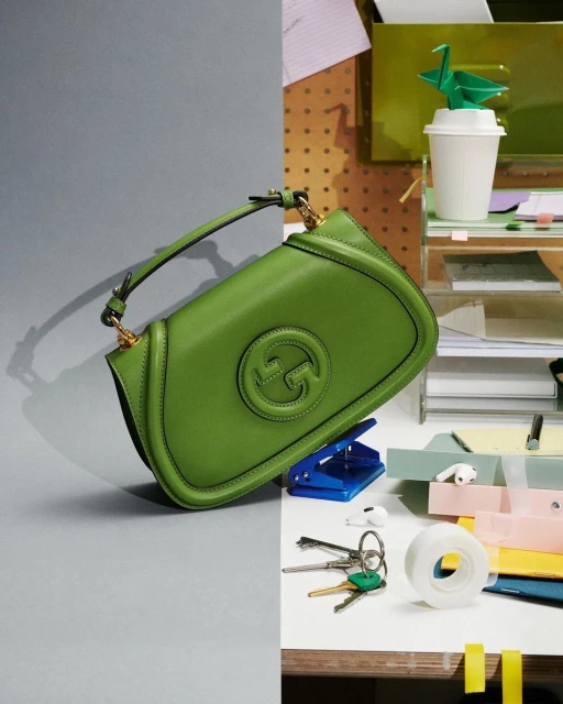 A green Gucci purse is presented against a split background of gray and a bright desk with office supplies.