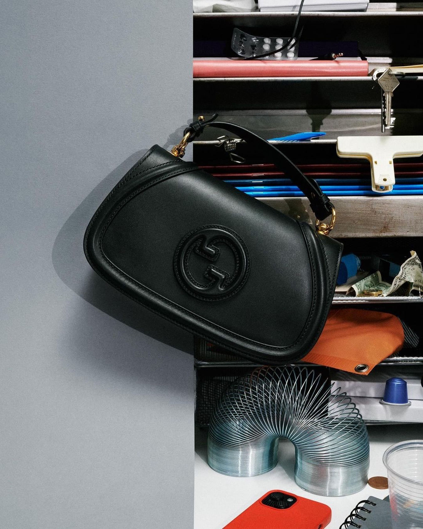 Black Gucci handbag on display against a gray wall next to various objects including a slinky toy and a red cell phone.