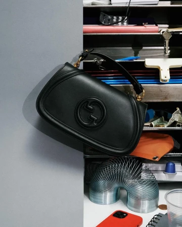 Black Gucci handbag on display against a gray wall next to various objects including a slinky toy and a red cell phone.