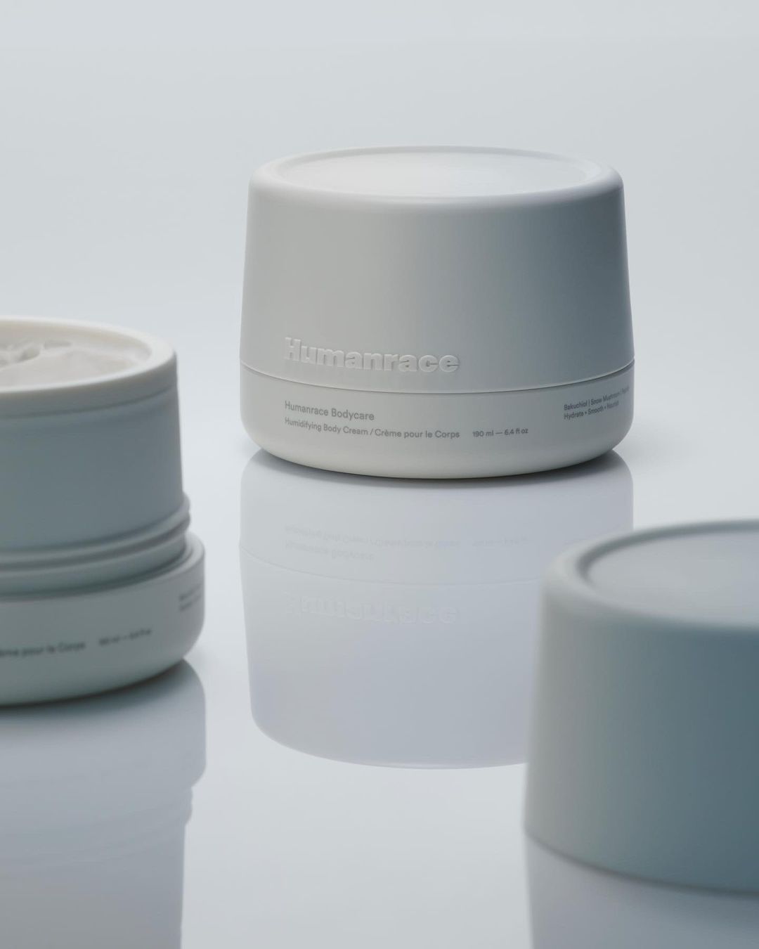 Three Humanrace skincare cream containers on a white reflective surface, featuring clean packaging and minimalist design for beauty product photography.