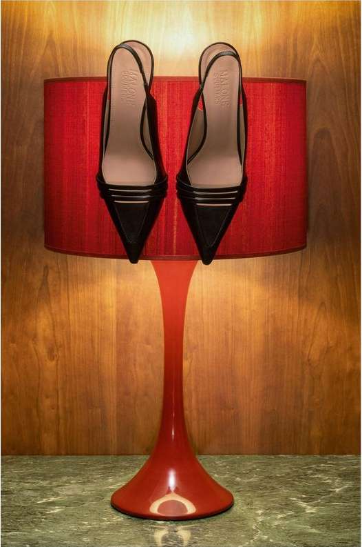 Pair of black slingback high-heeled shoes resting on a red tulip table lamp against a wood-paneled and marble-topped surface indoors.