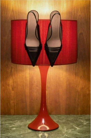Pair of black slingback high-heeled shoes resting on a red tulip table lamp against a wood-paneled and marble-topped surface indoors.