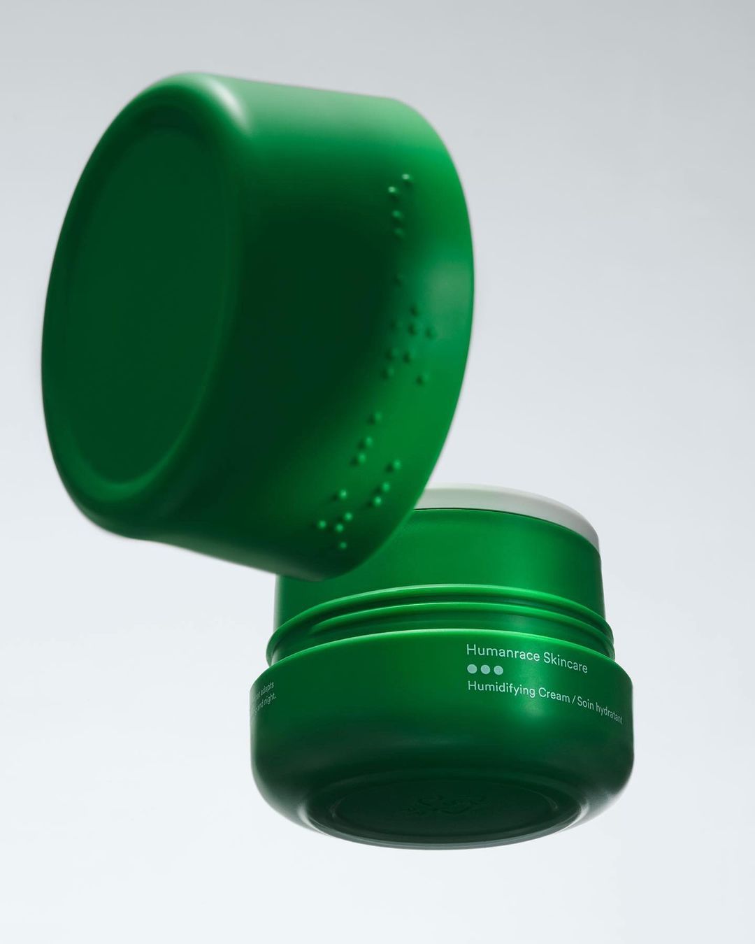 Close-up studio shot of a green Humanrace Humidifying Cream jar with lid slightly open, set against a neutral gradient backdrop.