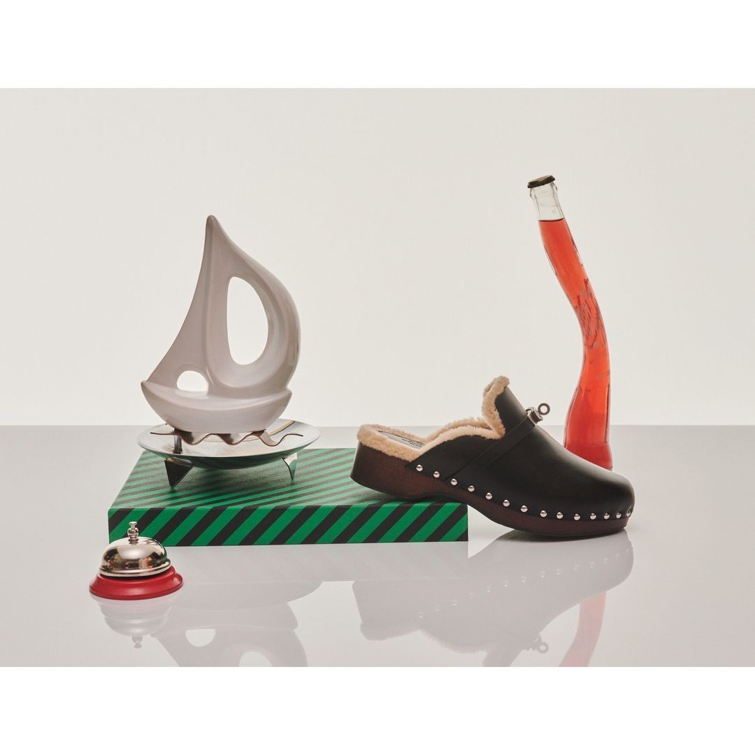 A brown clog is posed with a clear bottle with red liquid, a sailboat decoration, and a bell in a minimalist studio setting.