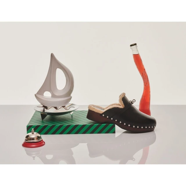 A brown clog is posed with a clear bottle with red liquid, a sailboat decoration, and a bell in a minimalist studio setting.