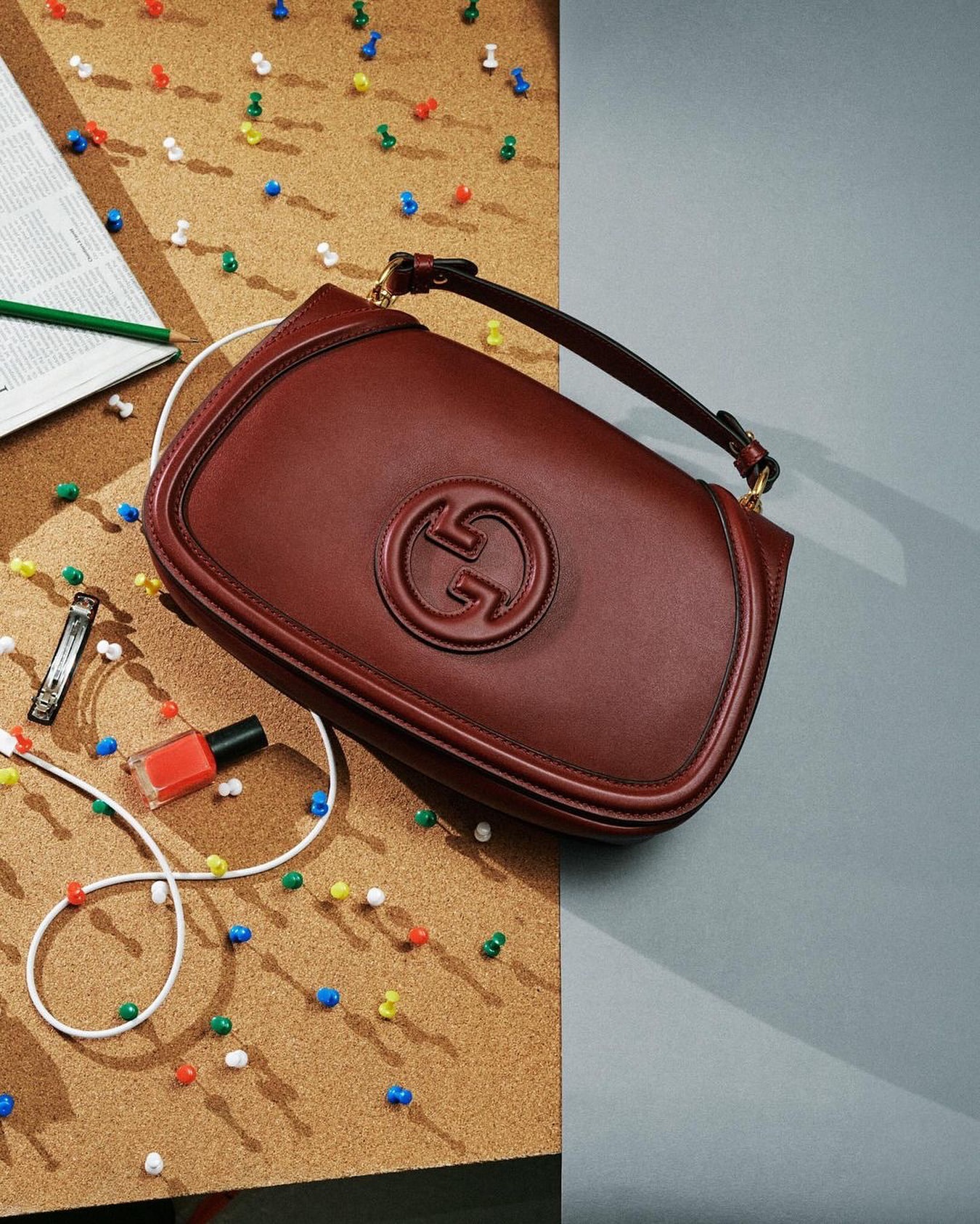 Close-up studio shot of a dark red Gucci handbag with a gold chain strap displayed on a cork board beside a nail polish bottle.