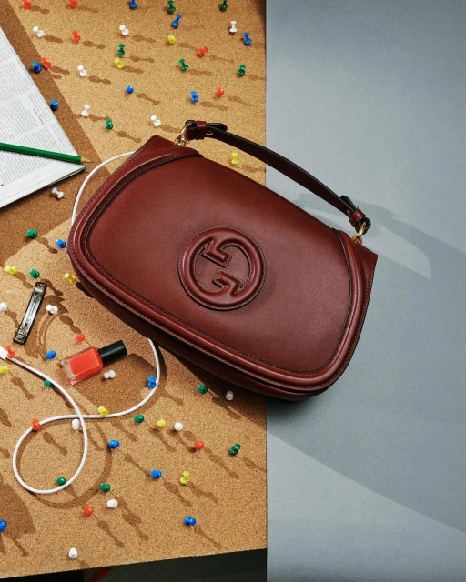 Close-up studio shot of a dark red Gucci handbag with a gold chain strap displayed on a cork board beside a nail polish bottle.