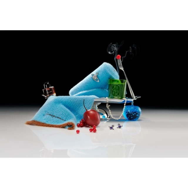 Still life composition featuring a blue fur accessory, candle, perfume bottle, cherry, and pearls against a black background.