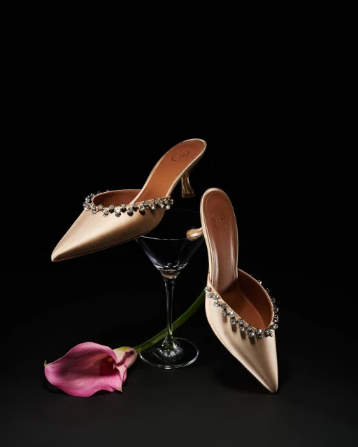 Elegant satin mule heels with crystal embellishments displayed with a calla lily and martini glass against a dark backdrop.