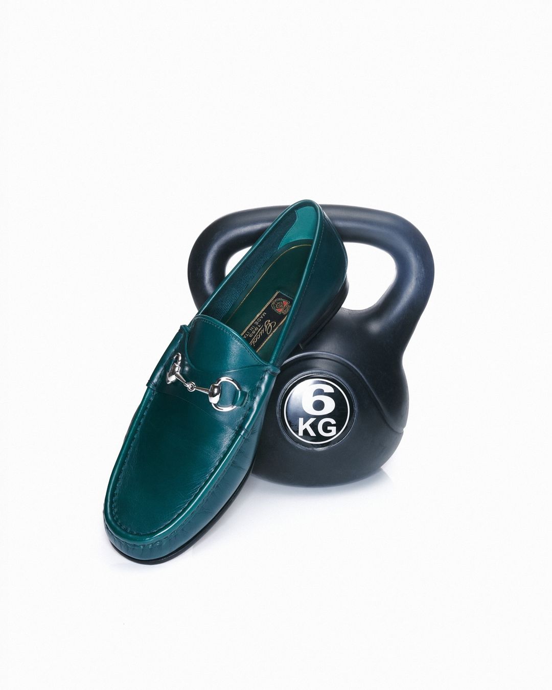 Elegant green leather loafer with a silver horse-bit detail alongside a black 6 kg kettlebell on a stark white backdrop, product still life.