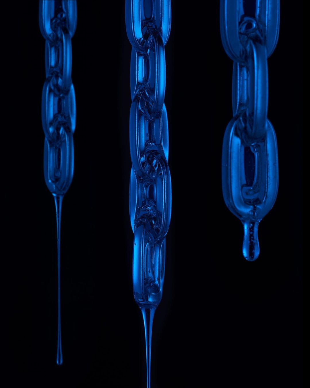 Three blue translucent chains with drips hang against a stark black background in a studio product shot with cool lighting.