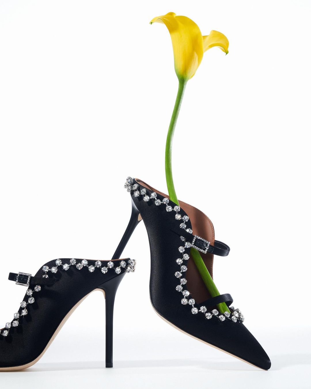Two black high-heel shoes decorated with clear gemstones are arranged on a white surface, one shoe holding a yellow calla lily.