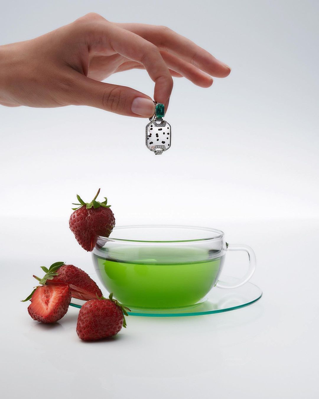 A hand holding a pendant over a glass cup with green liquid and strawberries on a white surface in a brightly lit studio setting.