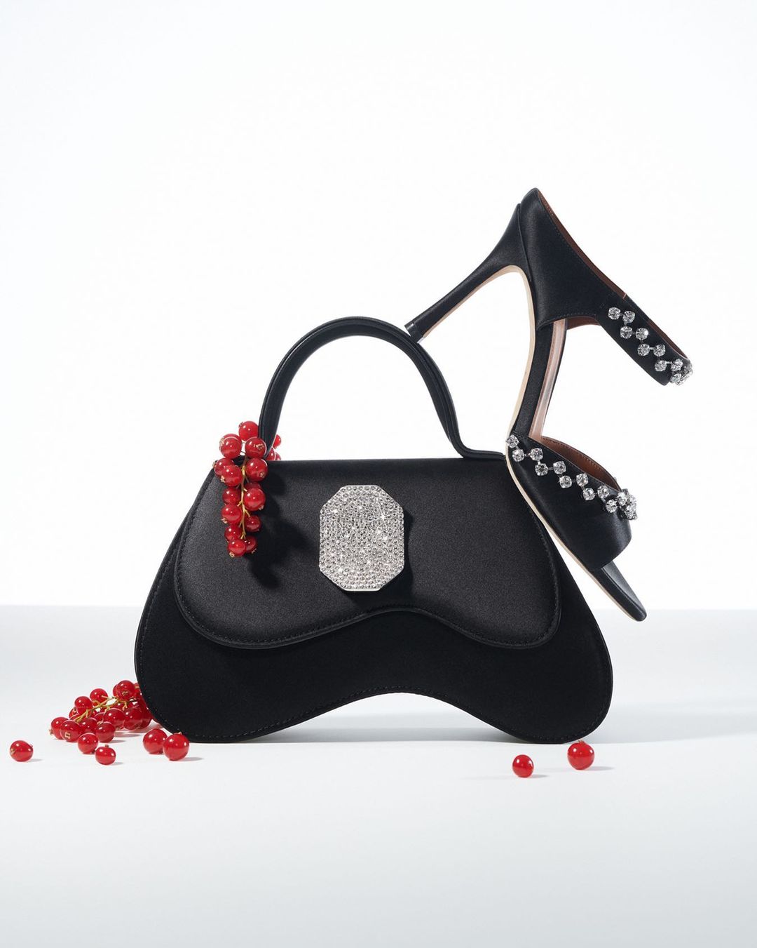 Black handbag with a rhinestone clasp next to a black high-heeled shoe adorned with rhinestones and a sprig of red currants.