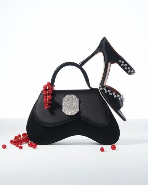 Black handbag with a rhinestone clasp next to a black high-heeled shoe adorned with rhinestones and a sprig of red currants.