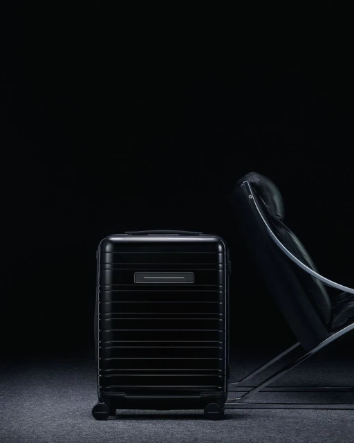 Sleek black carry-on luggage stands near a modern chair in a dim, studio-lit setting against a dark backdrop, emphasizing its design.