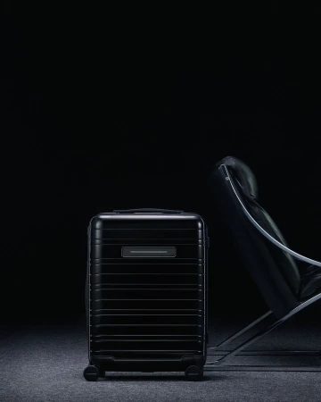 Sleek black carry-on luggage stands near a modern chair in a dim, studio-lit setting against a dark backdrop, emphasizing its design.
