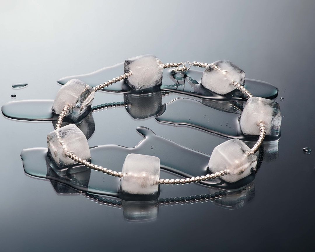 Still life of clear ice cube beads on a silver beaded string, reflected on a black surface, showing a pattern of cubes and water droplets.