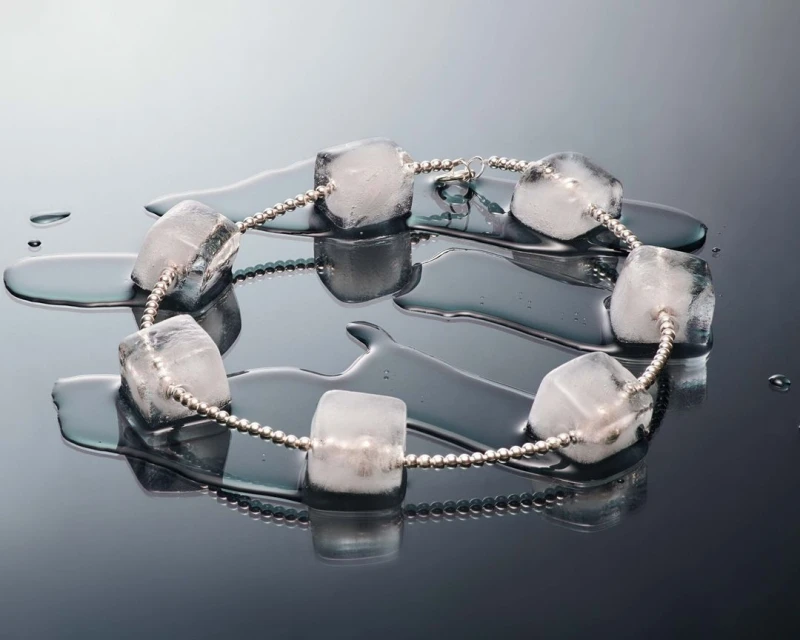 Still life of clear ice cube beads on a silver beaded string, reflected on a black surface, showing a pattern of cubes and water droplets.
